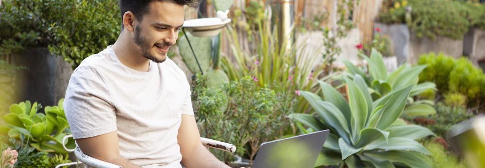 Eine lächelnde Person sitzt mit einem Laptop in einem Garten voller Pflanzen.